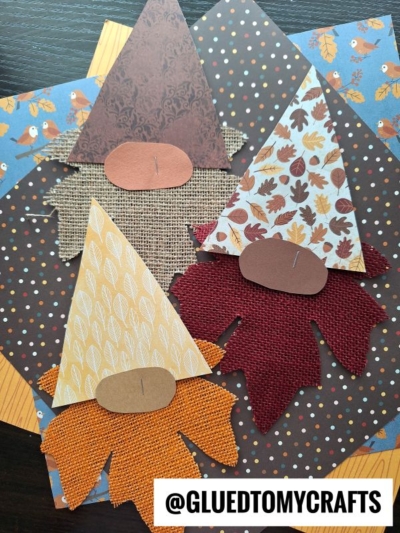 Three leaf gnome crafts made from patterned paper and textured fabric, featuring leaf-shaped beards and triangular hats, arranged on autumn-themed backgrounds. The handle @gluedtomycrafts appears at the bottom.