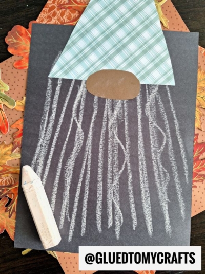 A fall-themed chalk art craft showing a gnome on black paper, featuring a plaid paper hat, brown oval nose, and a white chalk-drawn beard with a piece of chalk beside it. Autumn leaves decorate the background for this charming fall gnome.