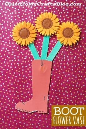 A paper boot flower vase holds three bright yellow sunflowers with green stems, set against a pink background with white and gold polka dots. Text reads BOOT FLOWER VASE.