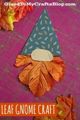 A charming leaf gnome craft featuring a handmade gnome with a leaf-patterned hat, beige oval nose, and orange artificial leaves as the beard, displayed on a pink textured background with extra leaves scattered around.