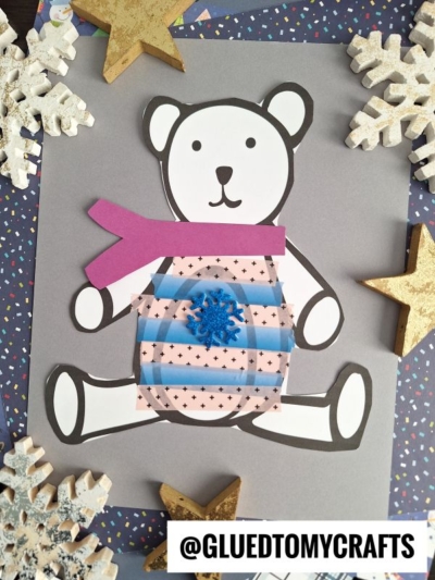 A washi tape bear in sweater style—a paper craft polar bear with a pink scarf, striped patterned belly, blue snowflake on its stomach, surrounded by wooden stars, snowflakes, and a navy blue border. @GLUEDTOMYCRAFTS text at the bottom.