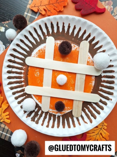 A fall-themed pie tic tac toe game on a paper plate with a painted orange center, popsicle sticks as the grid, and white and brown pom-poms as pieces. Surrounded by faux autumn leaves and a plaid napkin. @GLUEDTOMYCRAFTS.