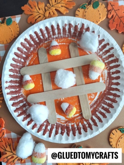 A paper plate decorated with brown paint transforms into a fun pie tic tac toe game board using craft sticks. Candy corn and cotton balls serve as game pieces, surrounded by festive felt pumpkin and leaf decorations.