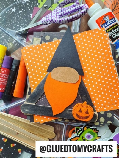 A popsicle stick Halloween gnome craft featuring a whimsical gnome with an orange hat and beard, a black triangle hat, and a small pumpkin, surrounded by craft supplies like glue sticks, popsicle sticks, and patterned paper.