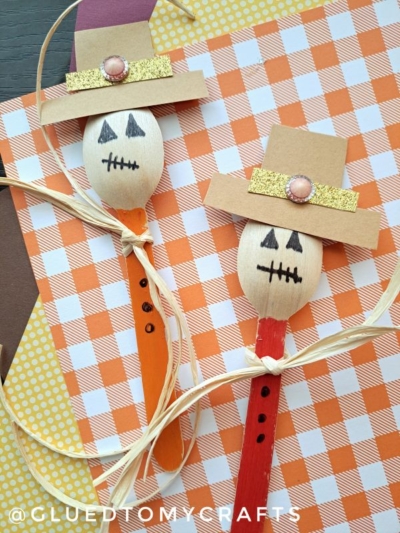 Two wooden spoon scarecrow figures feature drawn faces, paper hats, buttons, and raffia bows, displayed on orange and white checkered paper alongside other patterned sheets.