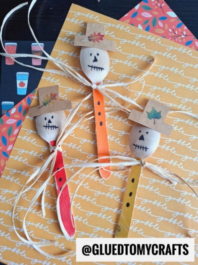 Three wooden spoon scarecrow faces with marker-drawn eyes and mouths, paper hats, and raffia bows are displayed on autumn-themed paper. @GLUEDTOMYCRAFTS is written at the bottom.