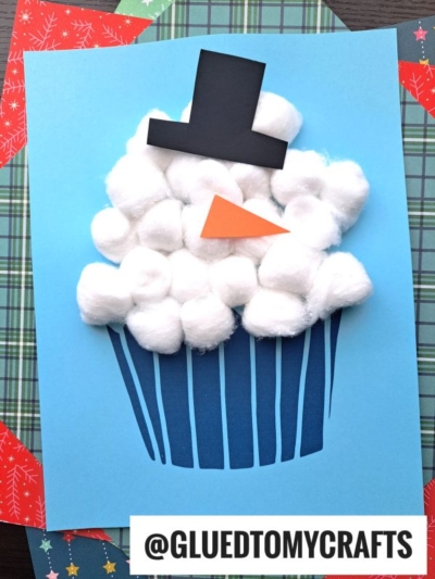 A cotton ball snowman cupcake craft featuring a white cotton ball top, black paper hat, and orange paper triangle nose, set on a blue background. Share your creations with @GLUEDTOMYCRAFTS!.