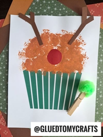 A festive reindeer cupcake craft made from paper and paint, featuring a brown cupcake top, green-striped wrapper, red nose, and brown antlers. Includes a clothespin with a green pom-pom. @GLUEDTOMYCRAFTS is written at the bottom.