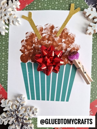 A holiday craft featuring a reindeer cupcake shape with brown sponge-painted “frosting,” gold antler cutouts, a large red shiny bow, and striped cupcake liner. Winter snowflake decorations and a clothespin with a pom pom are nearby.