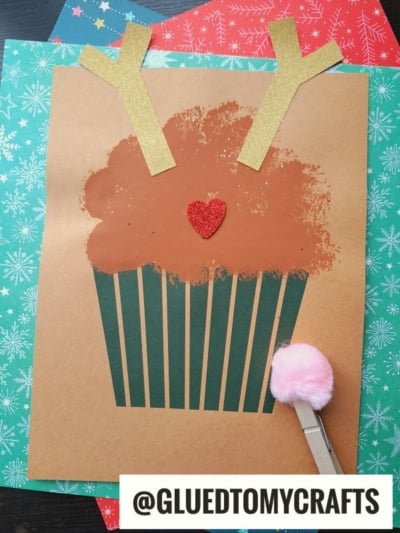 A charming reindeer cupcake craft shaped like a christmas cupcake, featuring brown paint, gold antlers, a red glitter heart nose, and a pink pom-pom on a clothespin, displayed on festive paper. @GLUEDTOMYCRAFTS is written at the bottom.