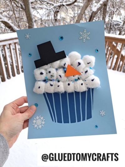 A hand holds a blue paper craft cotton ball snowman cupcake, featuring fluffy cotton frosting, a black hat, orange triangle nose, and blue sequins and snowflake stickers, set against a snowy outdoor background.