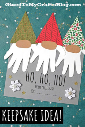 A festive craft features three santa gnomes made with patterned paper hats, oval noses, and white handprint cutout beards on a gray card that reads HO, HO, HO! MERRY CHRISTMAS! adorned with snowflakes and gold stars.