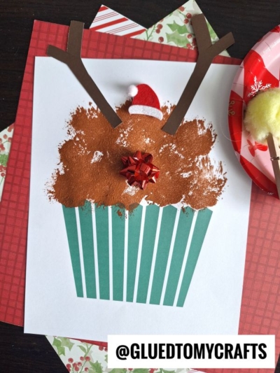 A festive reindeer cupcake craft features blue stripes, brown paint for fur, paper antlers, a mini Santa hat, and a red gift bow—all set on holiday paper with the handle @GLUEDTOMYCRAFTS. Perfect for Christmas cupcake inspiration!.