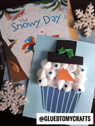A charming cotton ball snowman cupcake craft with blue hearts, a paper carrot nose, and a black top hat sits on blue paper beside the book The Snowy Day and two glittery snowflake ornaments. @gluedtomycrafts.