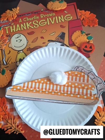 A Charlie Brown Thanksgiving book and a paper plate with a craft pie slice decorated with orange paper, washi tape pie accents, a cotton ball, and fall-themed craft supplies. @GLUEDTOMYCRAFTS is written at the bottom.