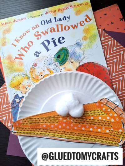 A children’s book titled I Know an Old Lady Who Swallowed a Pie sits on patterned paper. In front, there’s a paper plate with craft supplies—orange strips, googly eyes, cotton balls, and washi tape pie details—resembling a turkey. @gluedtomycrafts below.
