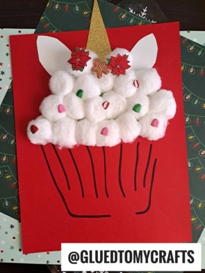 A cotton ball unicorn cupcake craft made with fluffy cotton balls, red and green gems, a shimmering gold horn, ears, red flowers, and a tiny gingerbread man on red paper, with @GLUEDTOMYCRAFTS printed at the bottom.
