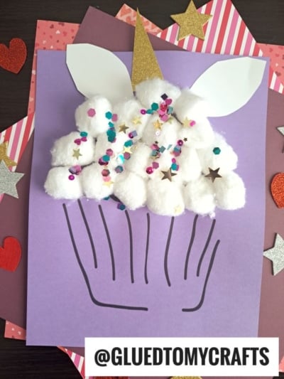 A cotton ball unicorn cupcake craft made with white cotton balls, sequins, gold star confetti, and cupcake lines on purple paper; features a gold horn, white ears, and colorful hearts in the background.