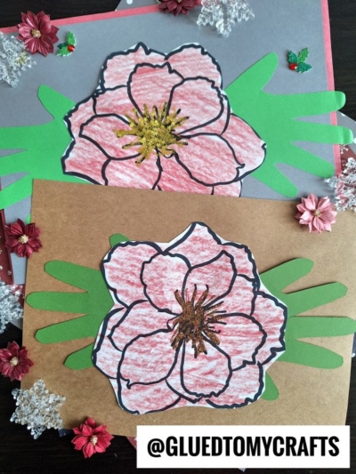 Two handmade Handprint Poinsettia Flower crafts with red and pink petals, green paper leaves, and glittery centers are displayed on brown and gray backgrounds, surrounded by small decorative flowers and snowflake ornaments.