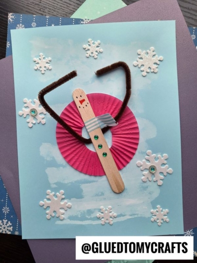 A winter craft featuring a popsicle stick snow tubing snowman with brown pipe cleaner arms, a gray bow tie, a pink cupcake liner, and felt snowflakes on a blue painted background. Text: @GLUEDTOMYCRAFTS.