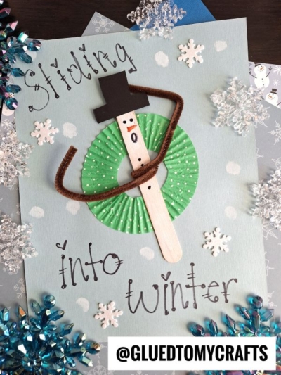 A handmade popsicle stick snow tubing snowman craft with a green cupcake liner, brown pipe cleaner scarf, black paper hat, on blue paper with “Sliding into winter” text and playful snowflake decorations.