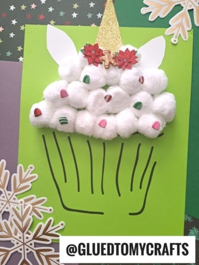 A cotton ball unicorn cupcake craft made of white cotton balls with mini decorations, paper ears, and a golden cone on green paper. Snowflakes and the @GLUEDTOMYCRAFTS label are also visible.