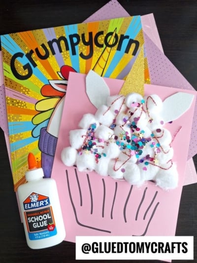 A children’s book titled Grumpycorn, a bottle of Elmer’s glue, and a pink cotton ball unicorn cupcake craft made with glitter and paper ears are arranged on a table. @GLUEDTOMYCRAFTS is visible at the bottom.
