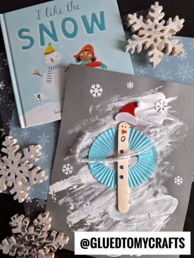 A children’s book titled “I Like the Snow” sits near a popsicle stick snowman craft, complete with a blue cupcake liner, red hat, and wooden snowflakes on gray paper. A tag reading @GLUEDTOMYCRAFTS is also shown.