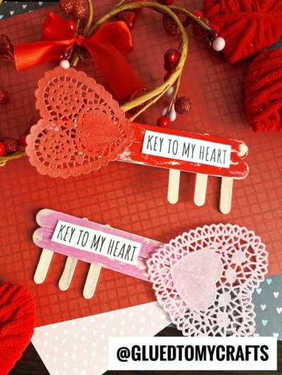Two handmade lock and key DIY popsicle sticks crafts, featuring colored popsicle stick keys and heart-shaped doilies, are displayed on a red background with paper hearts. The Instagram handle @GLUEDTOMYCRAFTS is at the bottom.
