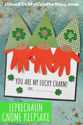 Three St. Patrick's leprechaun gnomes with handprint beards and green hats are displayed on a card reading, You are my lucky charm! Love, ______. Clovers and blue stars decorate the background. Text: Handprint Gnome Leprechaun Keepsake.