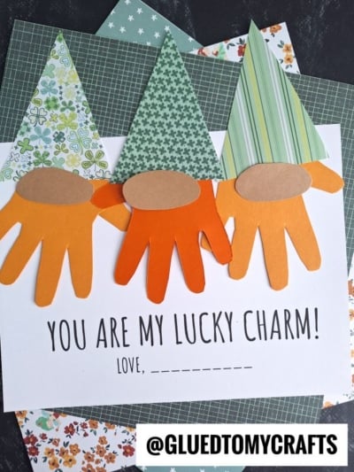 Three gnome leprechaun crafts, featuring patterned paper hats and orange handprint beards, are arranged above the text “You are my lucky charm! Love, _____” with the @GluedToMyCrafts watermark.