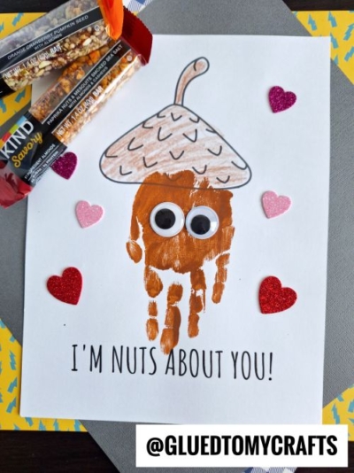 A cute handprint nuts about you card featuring an acorn with googly eyes, a brown cap, and pink and red glitter heart stickers. Below, “I'M NUTS ABOUT YOU!” is written, with a KIND snack bar placed on the left for a sweet keepsake.
