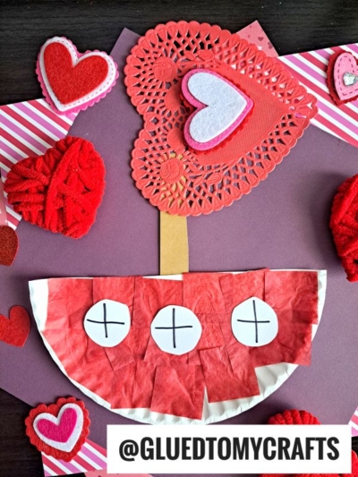 A Valentine’s Day craft featuring a heart-shaped tree made of red and pink paper, felt hearts, and a semi-circular base decorated with red tissue paper and white circles—perfect to pair with a paper plate love boat. The handle @gluedtomycrafts is at the bottom.