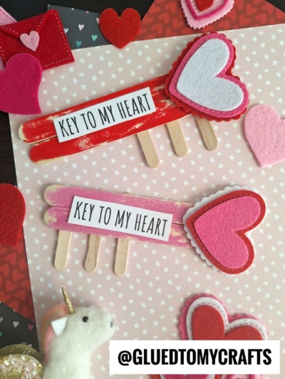 Two popsicle stick crafts shaped like arrows with heart tips and “Key to My Heart” labels are placed on heart-patterned paper, surrounded by felt hearts. A small unicorn figure sits in the corner—perfect for a lock and key DIY popsicle sticks project.
