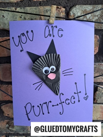 A handmade card on purple paper features a cupcake liner black cat face crafted from a black cupcake liner, googly eyes, pink nose, and drawn whiskers. The text reads, You are purr-fect! with @GLUEDTOMYCRAFTS at the bottom.