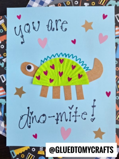 A card with a green cupcake liner dinosaur decorated with pink hearts, googly eye, and brown legs and tail. It reads You are dino-mite! in playful writing, with star and heart stickers on a blue background.