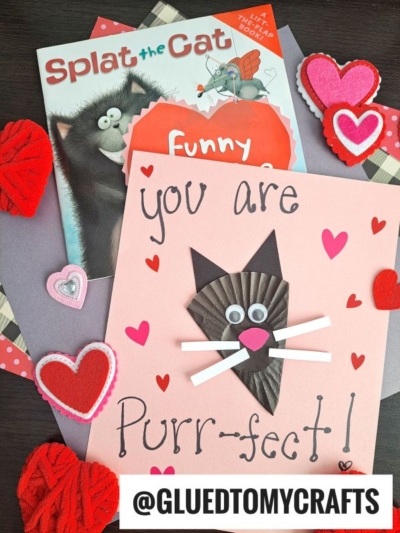 A handmade Valentine’s card featuring a cupcake liner black cat with googly eyes, whiskers, and “You are Purr-fect!” text, surrounded by heart decorations and Splat the Cat books on a dark surface.