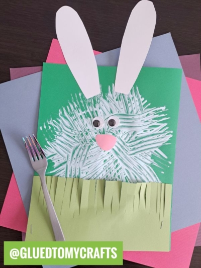 A handmade bunny fork painting craft made from green and pastel paper, with white fur textured using a fork, googly eyes, paper ears, a pink nose and mouth, and paper grass. @GLUEDTOMYCRAFTS is written in green at the bottom.