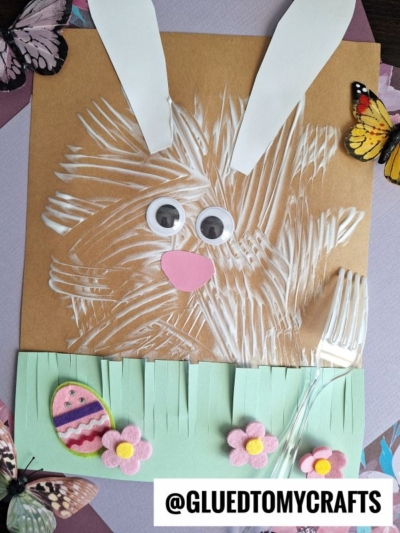 A fork painted bunny face made from white paint on brown paper, with paper ears, googly eyes, a pink nose, green paper grass, felt flowers, a decorated egg, butterflies, and a plastic fork lying nearby.