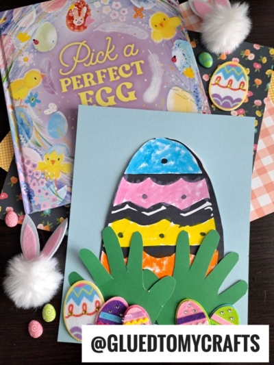 A colorful Easter-themed craft scene with a book titled Pick a Perfect Egg, handmade striped egg with handprint grass on blue paper, decorated eggs, bunny ears, and the Instagram handle @gluedtomycrafts.