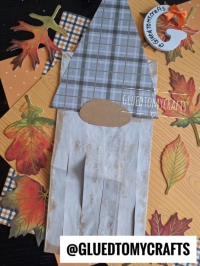 A fall-themed brown paper bag gnome craft featuring a plaid hat, surrounded by cut-out autumn leaves and orange patterned paper. @GLUEDTOMYCRAFTS is displayed on the image.