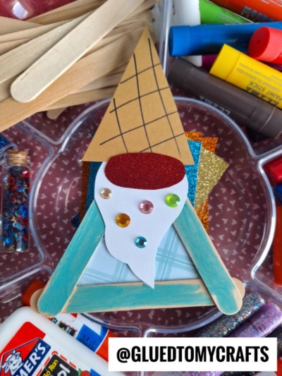 A handmade popsicle stick ice cream gnome crafted with colored paper, gems, and glitter, surrounded by craft supplies like glue, scissors, markers, and sequins. The @GLUEDTOMYCRAFTS handle is visible at the bottom.