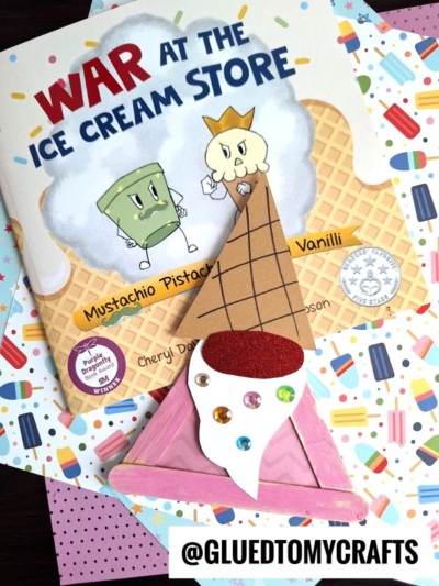 A children’s book titled “War at the Ice Cream Store” lies on colorful, patterned paper. Next to it is a handmade waffle cone gnome craft, sparkling with jewels and a bright red top like a cherry on an ice cream sundae.
