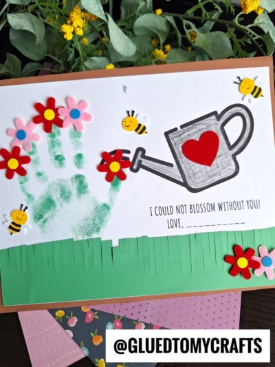 A handmade card features a green painted handprint, red felt flowers, a drawing of a watering can with a red heart, cartoon bees, and the sweet message: “I could not blossom without you! Love, ___.”.