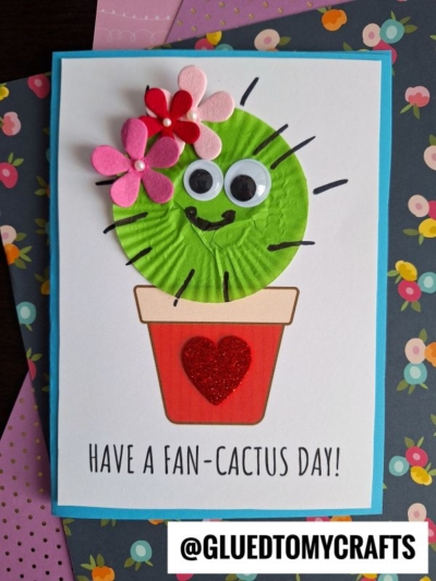 A handmade cupcake liner cactus card features a smiling cactus with googly eyes and paper flowers, set in a flowerpot with a red glitter heart. Text says "Have a fan-cactus day!" on a cheerful floral background.