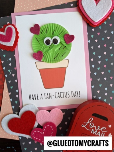 A handmade fan-cactus card features a green cactus with googly eyes and glittery heart shapes in a flowerpot, captioned “Have a fan-cactus day!” Surrounded by felt hearts and heart-patterned papers. @gluedtomycrafts.