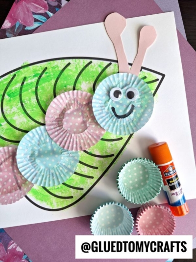 A cupcake liner caterpillar craft made with pastel polka-dot liners, googly eyes, and a drawn smile rests on a paper leaf outline. Nearby are a glue stick and extra liners. @GLUEDTOMYCRAFTS text is at the bottom.