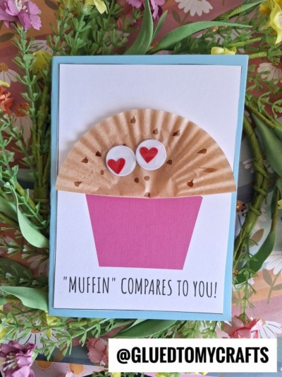 A handmade muffin card for mom features a muffin made from a brown cupcake liner and pink paper cup, with googly eyes and red hearts. Text reads: Muffin compares to you! Surrounded by artificial flowers.