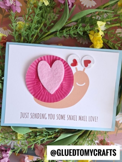 A handmade snail mail love card featuring a cupcake liner snail with a felt heart, drawn eyes, and hearts as antennae. “Just sending you some snail mail love!” is written on a charming floral background.