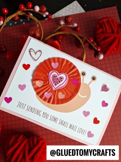 A handmade snail mail card featuring a cute cupcake liner snail and paper hearts. The card reads Just sending you some snail mail love! and is surrounded by red and pink hearts. The username @GLUEDTOMYCRAFTS is at the bottom.
